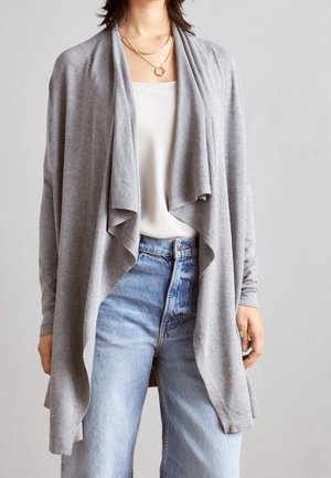 Femme portant un cardigan gris ample ouvert sur le devant, un haut blanc, un jean taille haute bleu, un collier en or et une bague en or, debout devant un fond uni.
