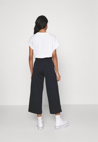 Bílý bavlněný tričko s krátkým rukávem v kombinaci s širokými námořnickými kalhotami. Model nosí bílé tenisky a ponožky ke kotníkům. Jednoduchý design.