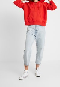 Sudadera roja con un bolsillo frontal y cuello con cordones, combinada con jeans cortos de lavado claro con dobladillos deshilachados y zapatillas blancas.