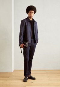 Veste navy avec fermeture éclair à l'avant, chemise noire, pantalon navy assorti et mocassins foncés. Tient un petit sac bandoulière noir avec une sangle.