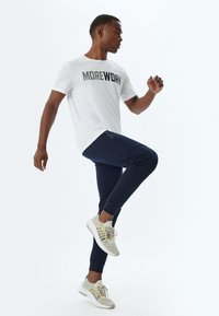 Witte sport T-shirt met "MOREWORK" in dikke letters gedrukt, gecombineerd met marineblauwe joggers met een ritszak en lichtgrijze sneakers.