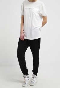 Maglietta bianca a maniche corte con tasca sul petto, abbinata a pantaloni neri a gamba affusolata e sneaker grigie. L'outfit presenta una vestibilità rilassata e tessuti morbidi.