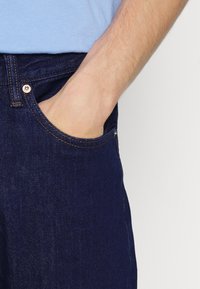 Jeans en denim bleu foncé avec une poche avant visible. Caractéristiques de surpiqûres orange et d'une fermeture à bouton en métal. Texture lisse et coupe classique.