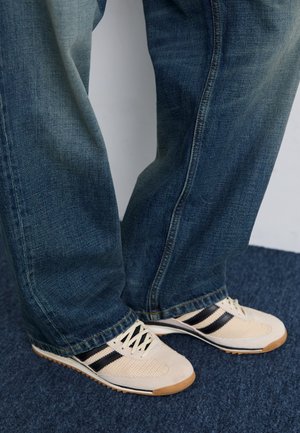 Person iført blå jeans og hvide sneakers med sorte striber, der står på en mørk tæppebelagt gulv.