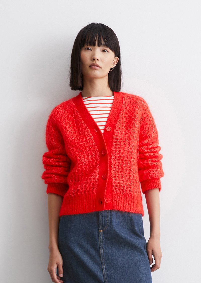 Cardigan rouge en tricot avec un motif texturé, doté d'un col en V, d'une fermeture à boutons et de manches retroussées, associé à un haut rayé et une jupe en denim.