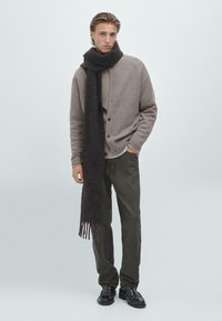 Jasnobrązowy kardigan z guzikami, beżowy sweter pod spodem, ciemnobrązowy szalik z frędzlami, ciemne spodnie i czarne mokasyny. Prosta, fakturowana tkanina.