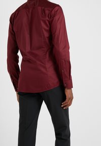 Camisa burdeos de manga larga con una textura suave, diseño ajustado y puños abotonados. Combinada con pantalones negros, que muestran una silueta elegante.