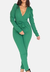 Grön jumpsuit med lång ärm, djup V-ringning, midjeband som dras åt och figurnära ben, tillverkad av slät tyg med svagt glans.