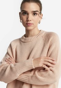Helle rosa Strickpullover mit geripptem Kragen und Ärmeln, der ein strukturiertes Muster und eine lässige Passform aufweist. Das Model hat die Arme verschränkt.
