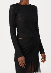 Svart långärmad topp i texturerat tyg, cropped design, scallop-kant och åtsittande ärmar, i kombination med en svart rynkad kjol.