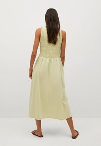 Robe sans manches de couleur jaune clair avec un corsage smocké et une jupe fluide, fabriquée en tissu doux, atteignant mi-mollet, portée avec des sandales plates.