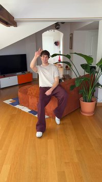 Oranžs kordurojas sofa ar apaļu formu, apkārt zaļi augi. Gaišas koka grīda, baltas sienas un melns TV skapis fonā.