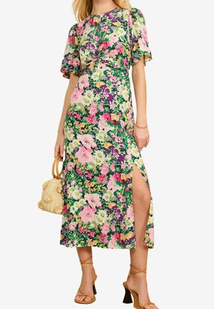 Kobieta ubrana w kwiecistą sukienkę do połowy łydki z bufkami na rękawach, przekręcanym detalem z przodu, bocznym rozcięciem, brązowymi sandałami na wysokim obcasie i trzymająca plecioną torebkę.