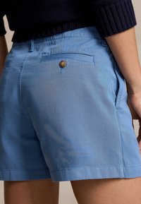 Ljust blå shorts av bomullstyg, med en bakficka med en mässingsknapp och en slät yta. Minimalt synligt sömnadsdetaljer.