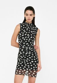 Schwarzes ärmelloses Kleid mit weißen Polka-Punkten, mit hochgeschlossenem Kragen und gerafftem Design, aus einem transparenten, dehnbaren Material gefertigt.