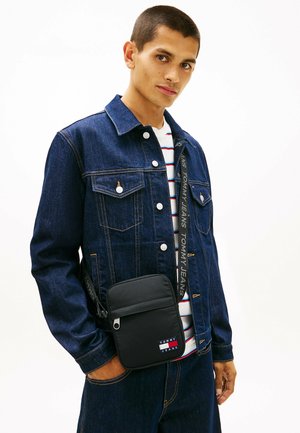 Jonge man die een donker denim jack en spijkerbroek draagt, een gestreept shirt, met een kleine zwarte Tommy Jeans schoudertas op een witte achtergrond.