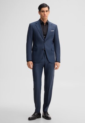 Man wearing a dark blue suit with a black shirt and black dress shoes standing against a plain light gray background.