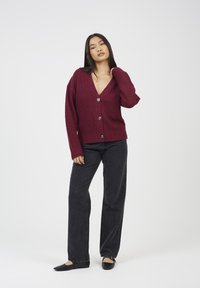 Kobieta w bordowym kardiganie zapinanym na guziki i czarnych szerokich spodniach, stojąca na jednolitym białym tle, trzymając się za kołnierz.