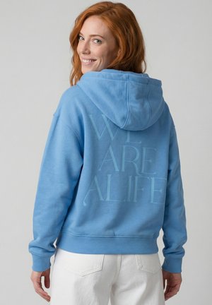 Rothaarige Frau lächelt über die Schulter, trägt einen blauen Kapuzenpullover mit dem Schriftzug "WE ARE ALIFE" auf dem Rücken und weiße Hose vor grauem Hintergrund.