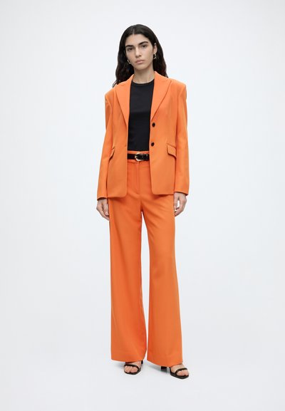 Femme aux longs cheveux foncés portant un tailleur orange avec un pantalon à jambes larges et un haut noir, debout sur un fond blanc.