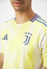 Gele voetbalshirt met grijze accenten, voorzien van een textuurpatroon, een embleem met drie sterren en contrasterende roze en blauwe schouderstrepen.