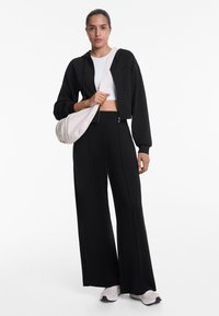 Haut à capuche noir zippé court au-dessus d'un crop top blanc, associé à un pantalon noir à taille haute et à jambes larges, ainsi qu'à des baskets légères. Porte un petit sac à bandoulière léger.