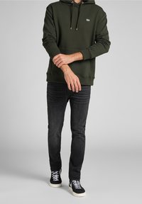 Man i mörkgrön Lee-hoodie, svarta jeans och svarta högstövelskor med vita snörning, står med armarna korsade framför sig.