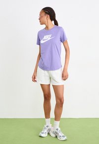 T-shirt Nike violet avec logo blanc, associé à un short blanc. Le modèle porte des baskets grises et roses, des chaussettes blanches et des lunettes roses. Sol vert.