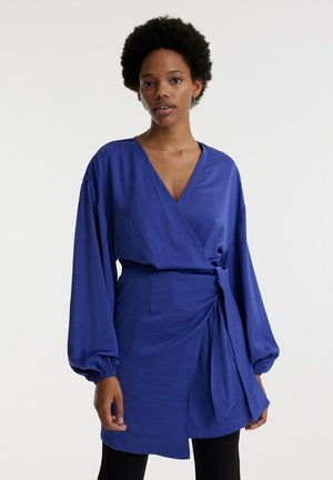 Blaue Wickelbluse mit langen, puffigen Ärmeln. Sie hat einen tiefen V-Ausschnitt und eine taillierte Schnürung. Hergestellt aus glattem Stoff mit einem leichten Glanz.