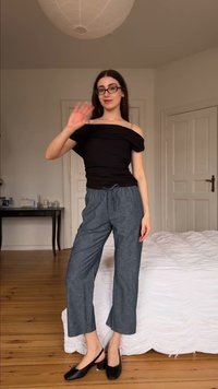 Femme portant des lunettes, un haut noir à épaules dénudées, un pantalon gris et des talons noirs, qui fait un signe de la main dans une chambre avec un sol en bois et un lit blanc.