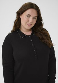 Polo à manches longues noir avec col, présentant des coutures contrastantes et six boutons sur le devant. Tissu doux et texturé.