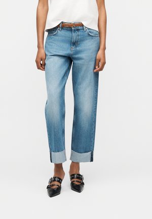 Persoană purtând blugi bleu deschis cu tiv întors, curea maro, bluză albă și pantofi negri cu vârf ascuțit și curele cu cataramă.