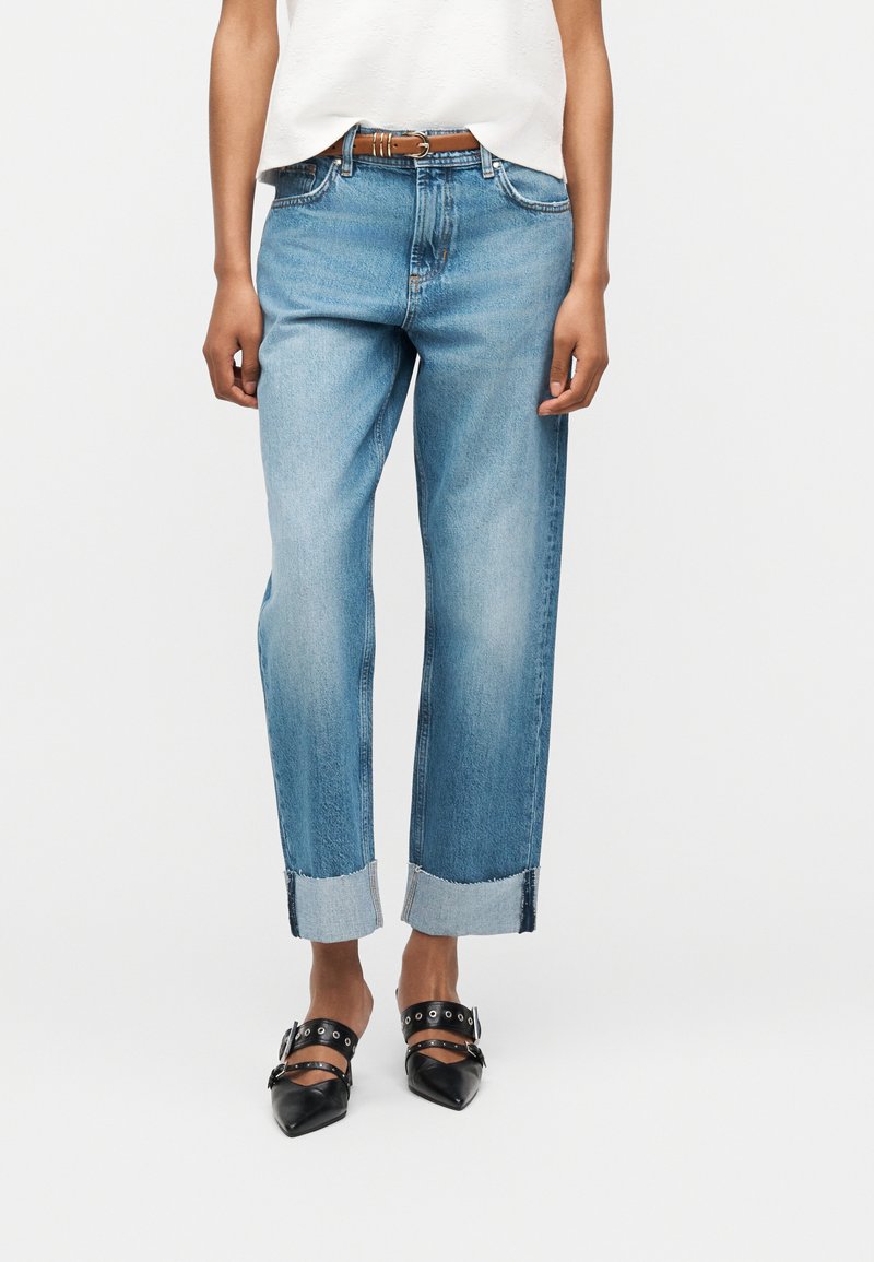 Άτομο που φοράει ανοιχτό μπλε τζιν με ρεβέρ, καφέ ζώνη, λευκή μπλούζα και μαύρα μυτερά παπούτσια με λουράκια και αγκράφες.