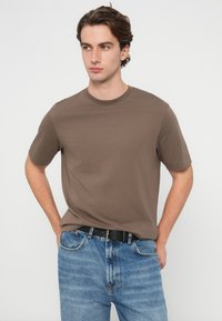 Rjava bombažna majica z okroglim izrezom in kratkimi rokavi, kombinirana s svetlo modrimi jeans hlačami. Tkanina je videti gladka in ima sproščen kroj.