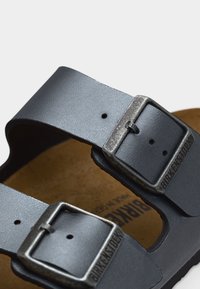 Δύο γκρι δερμάτινα λουριά σανδαλιών Birkenstock με μεταλλικές αγκράφες πάνω σε καφέ φελλό πατούρας.