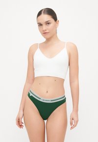 Balta tampri braletė su plonomis petnešėlėmis, derinama su žaliais aukštu liemeniu apatiniais, kurių baltas juostos raštas su "LACOSTE" logotipu.
