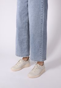 Helle beige Sneakers mit Schnürdesign, strukturierten Gummisohlen und einer klobigen, gerillten Plattform. Kombiniert mit hellblauen weit geschnittenen Jeans.