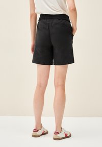 Pantaloni corti neri con vita elastica e due tasche posteriori. Abbinati a scarpe da ginnastica beige con dettagli rossi. Tessuto dalla consistenza liscia.