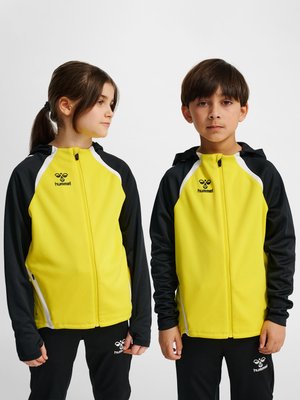 Dwoje dzieci w żółto-czarnych sportowych kurtkach Hummel oraz czarnych spodniach stoi obok siebie na jednolitym tle.