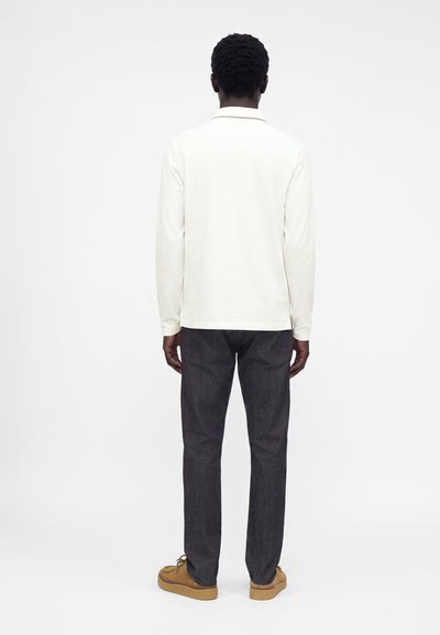 Homme debout de dos, portant une chemise blanche à manches longues, un jean foncé et des chaussures marron à lacets sur un fond uni.
