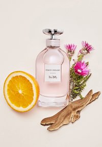Pink glass perfume bottle labeled "COACH DREAMS," accompanied by a sliced orange half, wood piece, and purple flowers on a light background.