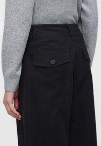 Gros plan d'une personne portant un pantalon noir avec des poches arrière boutonnées et un pull gris à manches longues, main reposant sur le côté.
