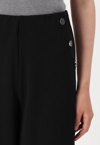 Zwarte, op maat gemaakte broeken met een gladde textuur zijn voorzien van decoratieve goudkleurige knopen aan de zijkant. Een aansluitend grijs geribbeld topje is gedeeltelijk zichtbaar.