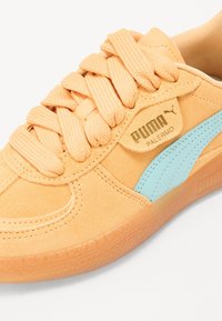 Zapatillas de ante naranja con acentos en azul claro, cordones texturizados, marca dorada y suela de goma translúcida. Diseño compacto y de perfil bajo.