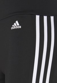Leggings sportivi neri con tre strisce bianche verticali sui lati e un piccolo logo Adidas bianco posizionato sopra le strisce.
