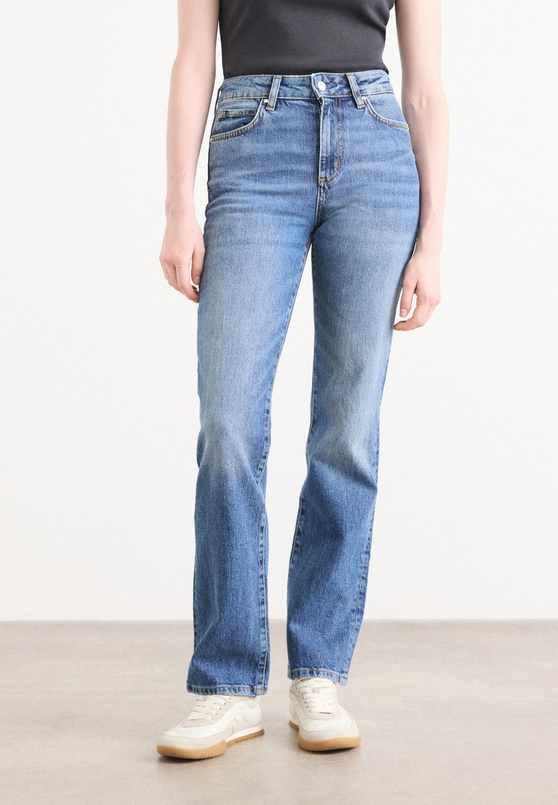 Lys blå denim jeans med høy midje, rett ben design, fem-lomme stil, og hvite og brune sneakers. Jeansen har en glatt tekstur og minimal detaljering.