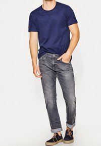 Camiseta de manga corta en color azul marino combinada con jeans de un gris claro, con los puños enrollados. El atuendo se completa con zapatillas oscuras azules y cordones marrones.