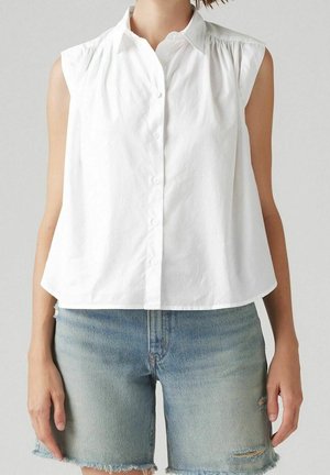 Chemisier blanc sans manches à boutons, associé à un short en denim bleu clair avec des ourlets usés et de légères déchirures.