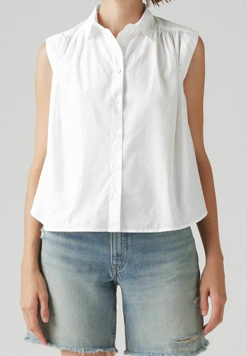 Chemisier blanc sans manches à boutons, associé à un short en denim bleu clair avec des ourlets usés et de légères déchirures.