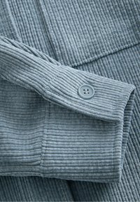 Blågråt fløjlstof med markant ribbet tekstur, med en knap på manchetten. Fokus på blødt materiale og strukturerede designdetaljer.